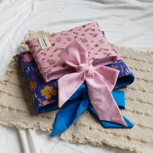 Peut inclure: Deux coussins décoratifs avec des motifs floraux et un nœud rose. Le coussin supérieur est rose avec un motif de feuilles noires, et le coussin inférieur est bleu avec des fleurs jaunes et roses. Un ruban bleu est noué autour des coussins.
