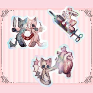 Könnte beinhalten: Set aus drei holografischen Aufklebern mit medizinischem Thema. Einer zeigt zwei katzenartige Figuren mit Messer und Schere, ein anderer ein Kaninchen mit einer Spritze und der letzte eine katzenartige Figur, die ein menschliches Herz hält. Die Aufkleber haben ein rosa und blaues Farbschema.