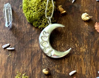 Puede incluir: Collar de plata en forma de media luna con un diseño detallado de las fases de la luna. El collar está sobre una superficie de madera con musgo y flores secas.
