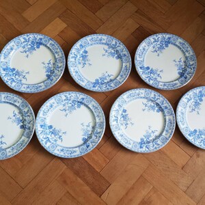 May include: Set of seven white ceramic dinner plates with a blue floral pattern. The plates have a scalloped edge and a raised rim.