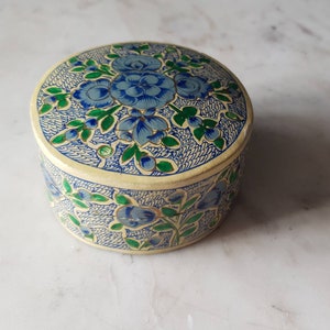 May include: A round, cream-colored trinket box with a floral design. The box is decorated with blue and green flowers and leaves.
