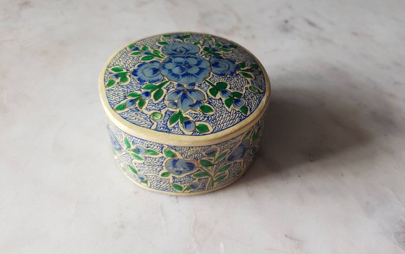 May include: A round, cream-colored trinket box with a floral design. The box is decorated with blue and green flowers and leaves.