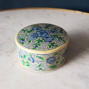 May include: A small, round, decorative box with a floral design. The box is made of paper and has a cream background with blue and green flowers.