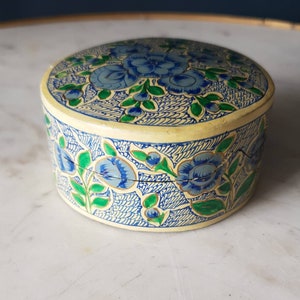 May include: A round, cream-colored box with a floral design. The box is decorated with blue and green flowers and leaves on a cream background. The box is made of paper mache.