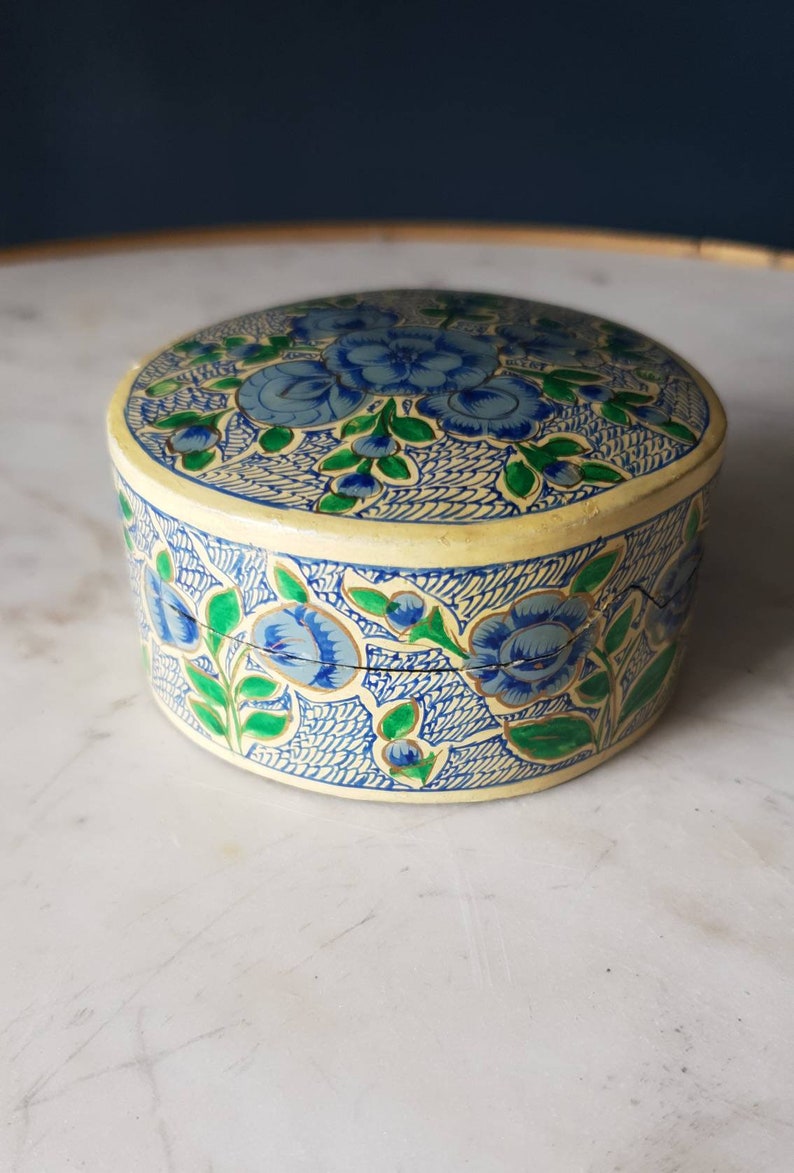 May include: A round, cream-colored box with a floral design. The box is decorated with blue and green flowers and leaves on a cream background. The box is made of paper mache.