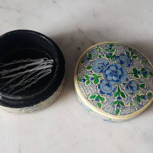 May include: A round, floral patterned box with a black interior. The box is open and contains a set of white bobby pins.