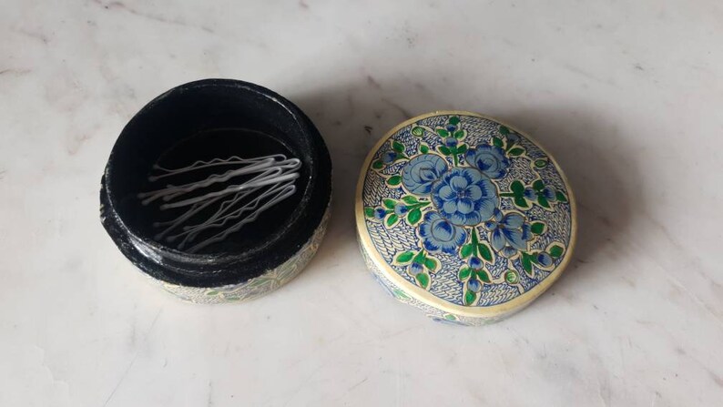 May include: A round, floral patterned box with a black interior. The box is open and contains a set of white bobby pins.