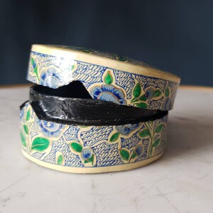 May include: A round, cream-colored box with a blue and green floral pattern. The box is slightly damaged and has a black interior.