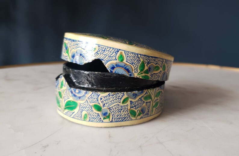 May include: A round, cream-colored box with a blue and green floral pattern. The box is slightly damaged and has a black interior.