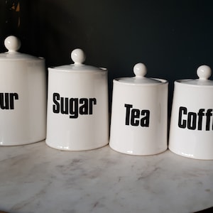 May include: Four white ceramic canisters with black labels. The labels read "Flour", "Sugar", "Tea", and "Coffee".