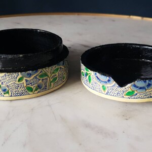 May include: A round, decorative box with a black interior and a floral pattern on the exterior. The pattern features blue flowers, green leaves, and a white background.