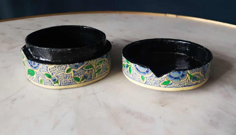 May include: A round, decorative box with a black interior and a floral pattern on the exterior. The pattern features blue flowers, green leaves, and a white background.