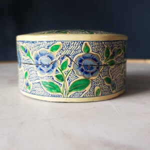 May include: A round, cream-colored trinket box with a floral design. The box is decorated with blue and green flowers and leaves.