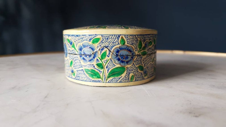 May include: A round, cream-colored trinket box with a floral design. The box is decorated with blue and green flowers and leaves.