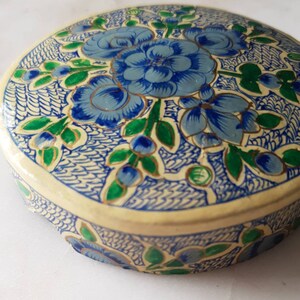 May include: A round, cream-colored box with a blue and green floral design. The box is decorated with intricate patterns and has a gold trim.