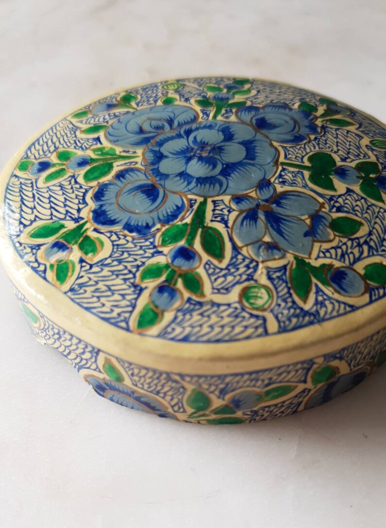 May include: A round, cream-colored box with a blue and green floral design. The box is decorated with intricate patterns and has a gold trim.