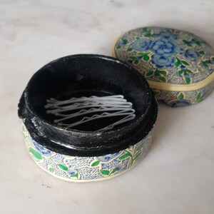May include: A small, round, floral patterned box with a black lid. The lid is open and contains a stack of white hair clips.