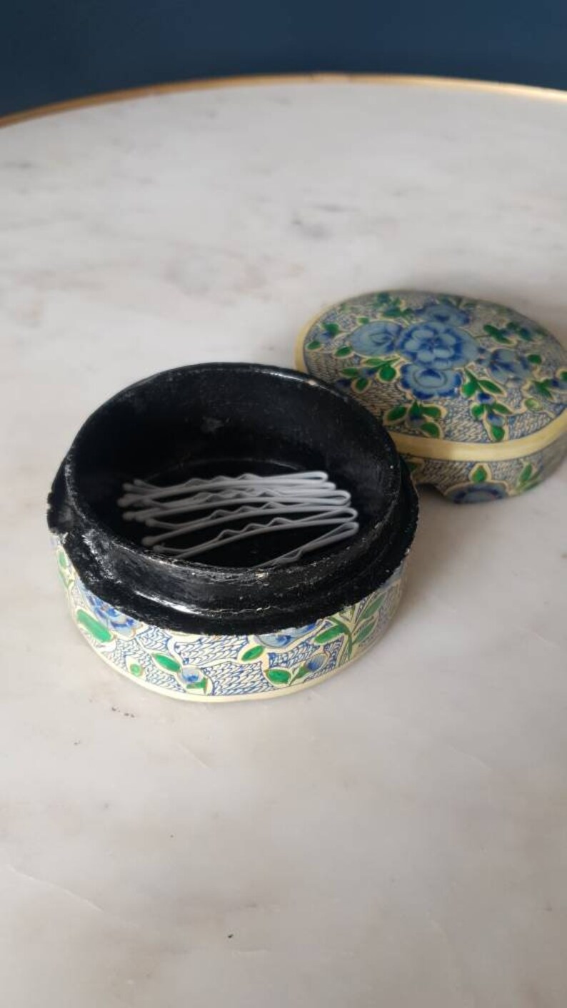 May include: A small, round, floral patterned box with a black lid. The lid is open and contains a stack of white hair clips.