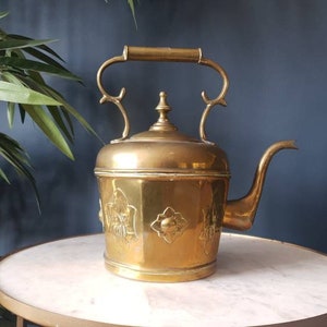 May include: A vintage brass teapot with an ornate design and a decorative handle. The teapot has a rounded body and a spout on the side. The teapot is sitting on a white marble surface.