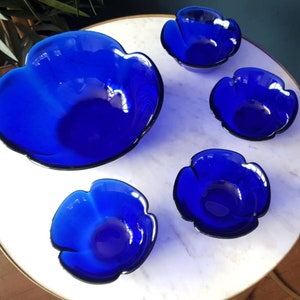 May include: Set of five cobalt blue glass bowls with a flower-shaped design. The bowls are arranged on a white marble surface.
