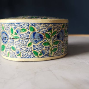 May include: A small, round, cream-colored box with a blue and green floral design. The box is made of paper mache and has a hinged lid.