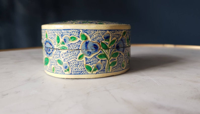 May include: A small, round, cream-colored box with a blue and green floral design. The box is made of paper mache and has a hinged lid.