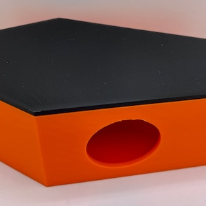 May include: An orange and black coffin-shaped box with a rounded opening. The box has a black, textured lid and orange sides. The opening is a large oval shape. The box is likely a pet house or a decorative storage container.
