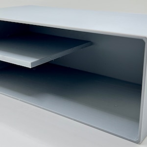 Puede incluir: Caja de almacenamiento rectangular gris claro con un estante interno. La caja tiene un agujero redondo en un lado. Fabricada con un material sólido y con un acabado liso y mate. El estante está posicionado horizontalmente.