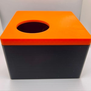 Può includere: Scatola di fazzoletti bicolore di forma quadrata. La parte superiore è arancione brillante con un'apertura circolare e la base è nera. La scatola è realizzata in un materiale solido e ha un design pulito e moderno.
