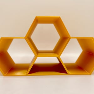 Peut inclure: Étagère jaune en forme de nid d'abeille avec trois compartiments hexagonaux. L'étagère est faite d'un matériau solide et est conçue pour être fixée au mur. L'étagère est de couleur jaune vif.