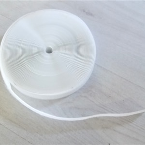 May include: A white roll of hook and loop tape, also known as Velcro, is shown on a light wood surface. The tape is rolled up and the end of the tape is unrolled and laying on the surface.