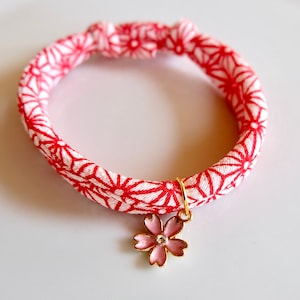 Peut inclure: Un bracelet &agrave; motif rouge et blanc avec un charm dor&eacute; en forme de fleur rose. Le bracelet est fait d'un tissu avec un motif g&eacute;om&eacute;trique r&eacute;p&eacute;titif. Le charm de la fleur a un petit bijou clair au centre.