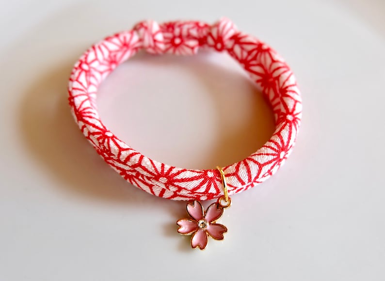 Peut inclure: Un bracelet &agrave; motif rouge et blanc avec un charm dor&eacute; en forme de fleur rose. Le bracelet est fait d'un tissu avec un motif g&eacute;om&eacute;trique r&eacute;p&eacute;titif. Le charm de la fleur a un petit bijou clair au centre.