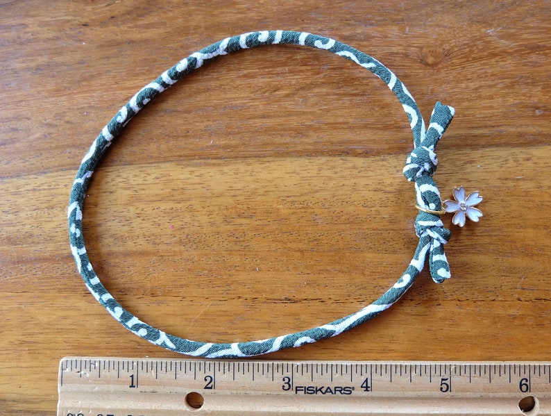 Peut inclure: Un collier en tissu &agrave; motifs verts et blancs avec un petit charme en forme de fleur. Le collier mesure environ 25 cm de diam&egrave;tre. Le charme est une petite fleur rose clair avec un anneau dor&eacute;.