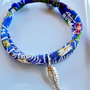 May include: A blue fabric bracelet with a silver fish charm. The bracelet is made of a patterned fabric with blue, green, and gold accents. The fish charm is detailed with scales and fins.