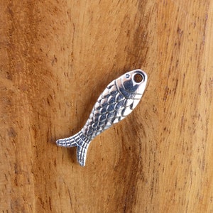 Peut inclure: Un charm en forme de poisson de couleur argent&eacute;e avec un motif d'&eacute;cailles d&eacute;taill&eacute;. Le charm a un petit trou en haut pour l'enfilage et une nageoire caudale d&eacute;taill&eacute;e. Le poisson est pr&eacute;sent&eacute; sur une surface en bois.