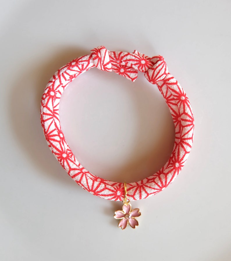 Peut inclure: Un collier pour animaux de compagnie &agrave; motif rouge et blanc, orn&eacute; d'un charm en forme de fleur de cerisier dor&eacute;. Le collier est nou&eacute; en haut.