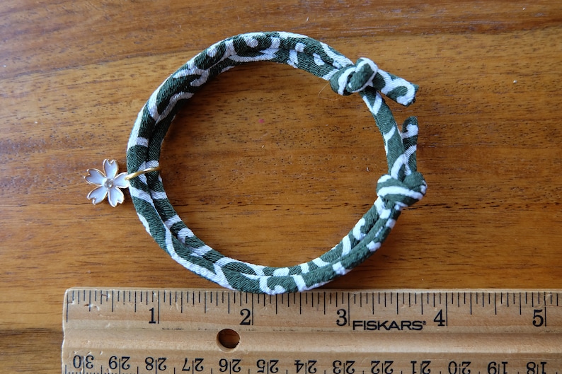 Peut inclure: Un bracelet en tissu &agrave; motifs verts et blancs avec un charm en forme de fleur blanche. Le bracelet est nou&eacute; aux extr&eacute;mit&eacute;s et poss&egrave;de un anneau dor&eacute; retenant le charm. Le bracelet mesure environ 12,7 cm de long.
