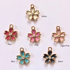 Peut inclure: Une collection de breloques en forme de fleur de diff&eacute;rentes couleurs, dont blanc, rouge, rose, rose clair, bleu et noir. Chaque breloque a une bordure dor&eacute;e et un petit bijou transparent au centre. Les couleurs sont &eacute;tiquet&eacute;es sous chaque breloque.