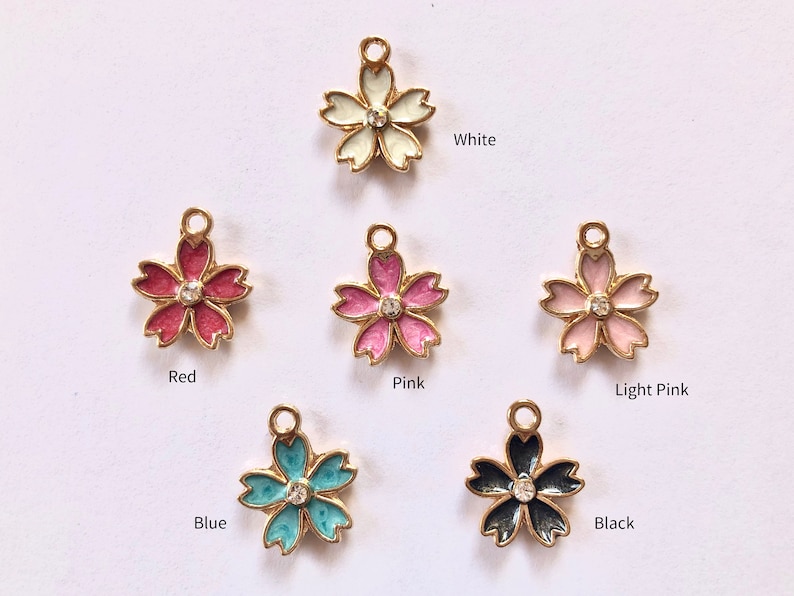 Peut inclure: Une collection de breloques en forme de fleur de diff&eacute;rentes couleurs, dont blanc, rouge, rose, rose clair, bleu et noir. Chaque breloque a une bordure dor&eacute;e et un petit bijou transparent au centre. Les couleurs sont &eacute;tiquet&eacute;es sous chaque breloque.