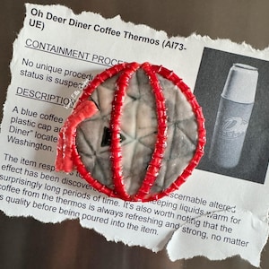 May include: A red, circular object with a textured surface and a red, segmented border. The object is on a torn piece of paper with text, including "Oh Deer Diner Coffee Thermos" and a drawing of a thermos.