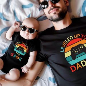 May include: A black t-shirt with the text "Leveled Up To Daddy" in a retro style with a sunset background and a video game controller graphic. A baby is wearing a black onesie with the text "Player 2 Has Entered The Game" in a retro style with a video game controller graphic.