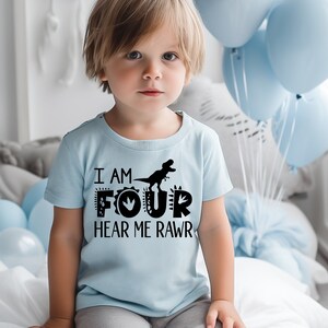 Peut inclure: Un t-shirt bleu clair avec un motif de dinosaure noir et le texte "I am Four Hear Me Rawr".