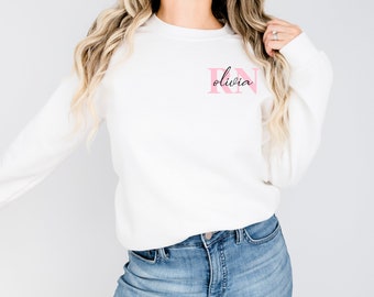 May include: White sweatshirt with pink and black lettering that says "Olivia RN".