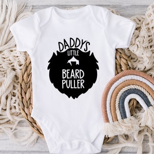 May include: White baby onesie with black text that reads "Daddy's Little Beard Puller" with a black silhouette of a beard.