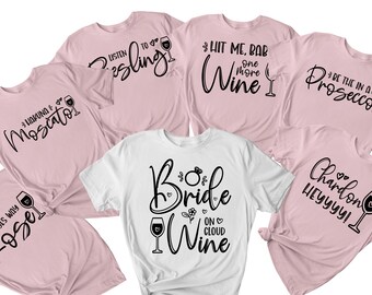 May include: A selection of pink and white t-shirts with wine-related slogans in black script. Slogans include "Listen to Riesling," "Hit Me, Bab, One More Wine," and "Bride on Cloud Wine." Each shirt has a wine glass illustration.