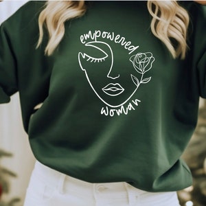 Könnte beinhalten: Ein dunkelgrüner Pullover mit einem weißen Grafikdesign eines Frauenkopfes mit einer Rose. Der Text "empowered woman" ist um die Grafik herum geschrieben.