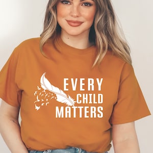 May include: A mustard yellow t-shirt with a white graphic of a feather and birds flying away. The text "EVERY CHILD MATTERS" is printed on the shirt.