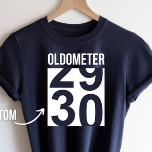 May include: A navy blue t-shirt with white text that reads "OLDOMETER 29 30". The text is in a bold, sans-serif font.