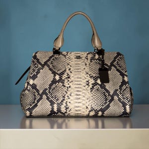 May include: A structured handbag featuring a grey and black snakeskin pattern with a central vertical stripe. The bag includes two light grey top handles, a gold-toned Coach logo on the front, and a black leather luggage tag hanging from the side.